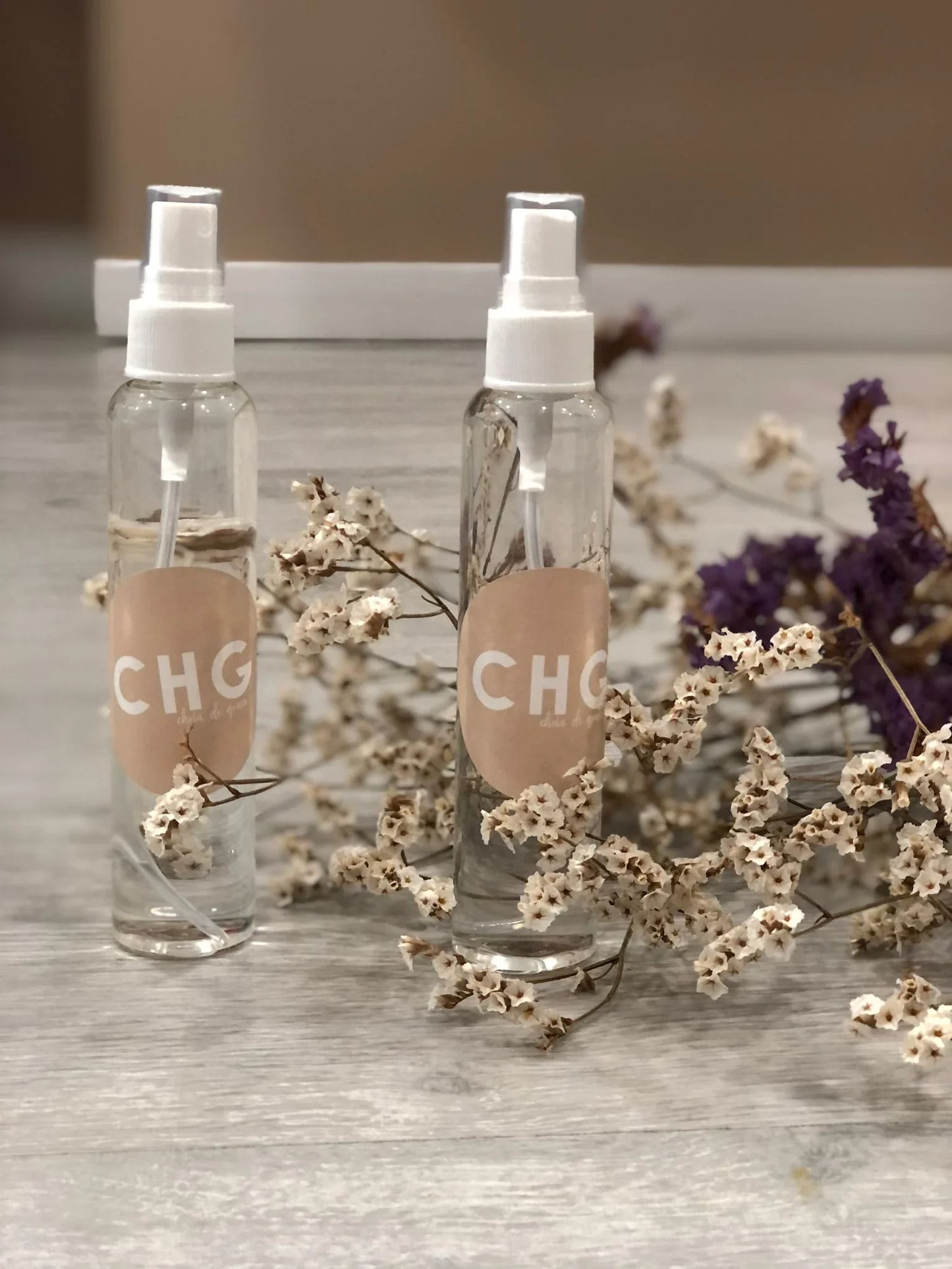 Dois frascos de spray CHG sobre mesa de madeira, decorados com flores secas, estilo elegante.