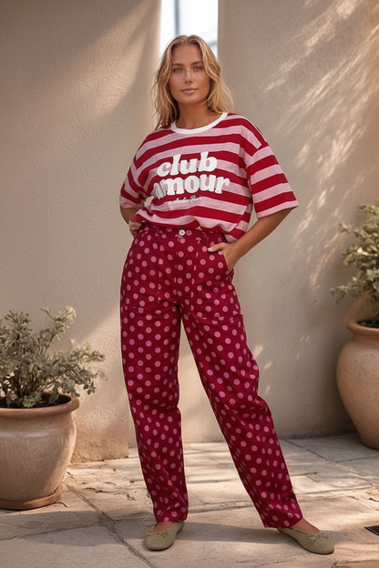 Modelo de cabelos claros vestindo calças Dotty vermelhas com padrão de bolas rosa, corte reto, bolsos laterais e elástico no cós combinadas com t-shirt às riscas.