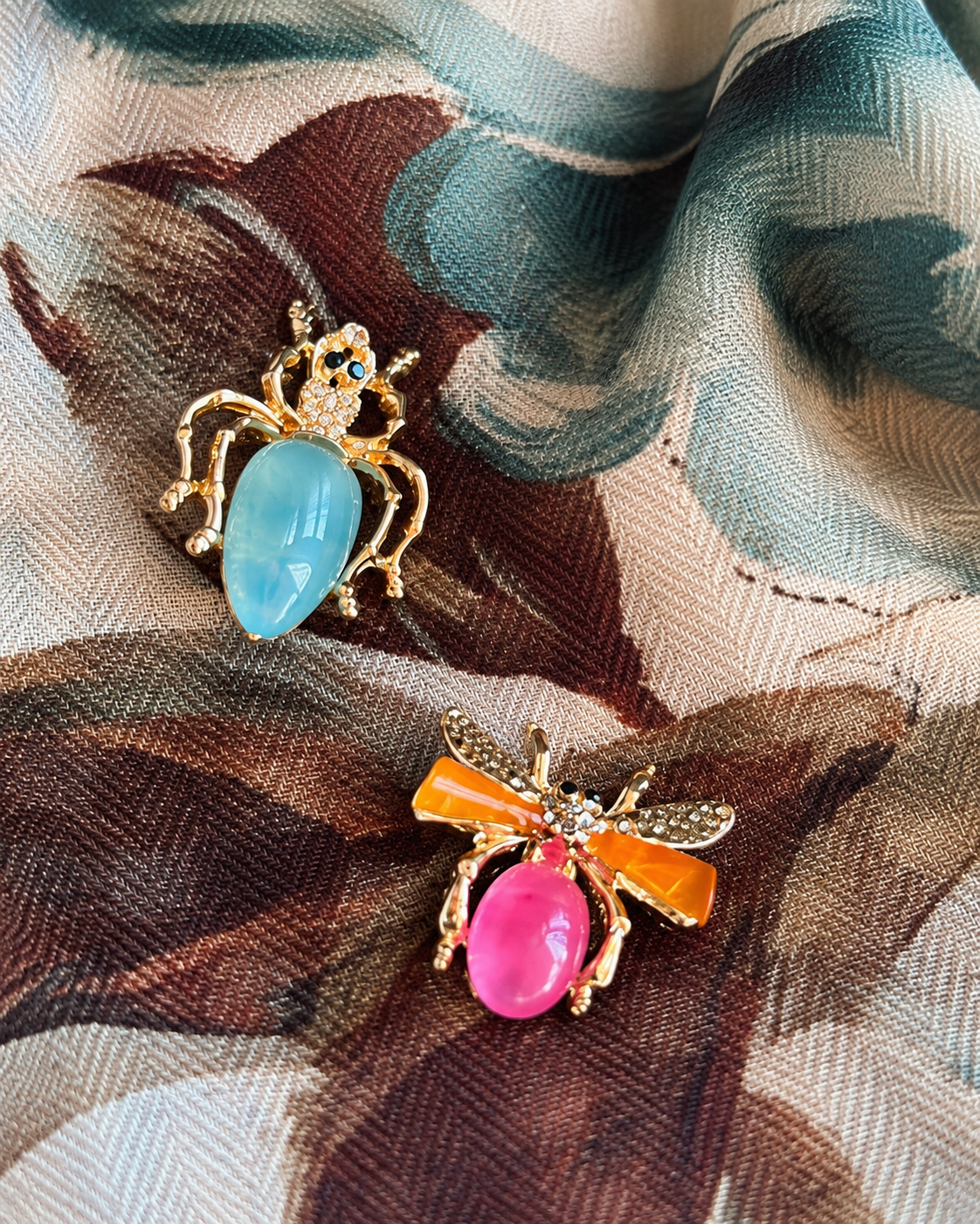 Conjunto de duas pregadeiras em forma de insetos, com acabamento dourado, pedras em azul-claro e rosa-fúcsia e detalhes brilhantes, sobre um lenço estampado.