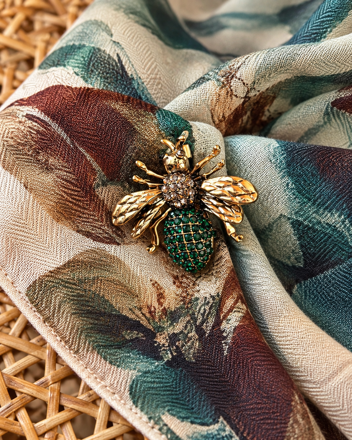 Pregadeira em forma de abelha, com acabamento dourado, asas trabalhadas, detalhes brilhantes e corpo decorado com pedras verdes, sobre um lenço estampado.