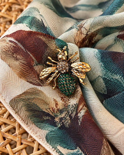Pregadeira em forma de abelha, com acabamento dourado, asas trabalhadas, detalhes brilhantes e corpo decorado com pedras verdes, sobre um lenço estampado.