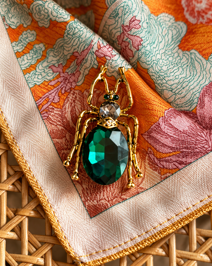 Pregadeira em forma de inseto, com acabamento dourado, corpo em pedra facetada verde-esmeralda e detalhes brilhantes, sobre um lenço estampado em tons de laranja e rosa.