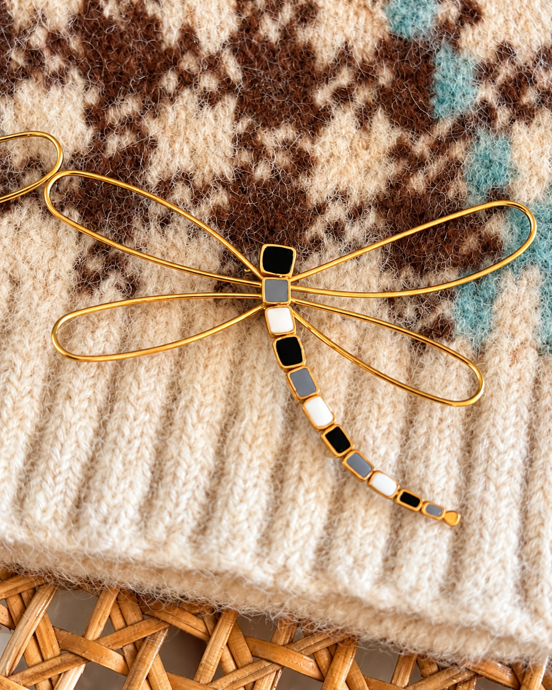 Pregadeira em forma de libélula, com asas delicadas em metal dourado e corpo com detalhes em preto, branco e cinzento, sobre uma malha bege estampada.