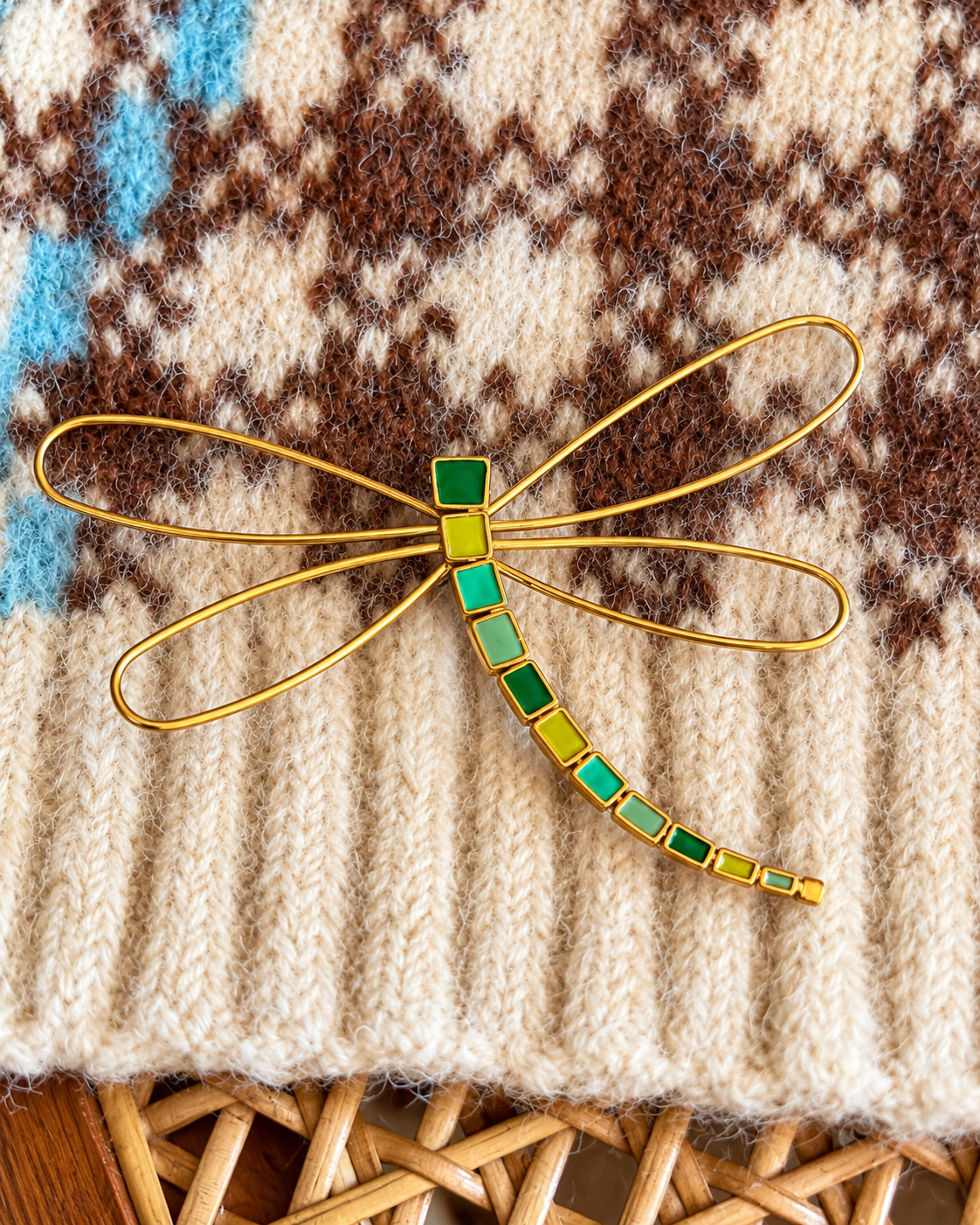 Pregadeira em forma de libélula, com asas delicadas em metal dourado e corpo decorado em tons de verde, turquesa e verde-lima, sobre uma malha bege estampada.