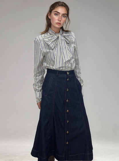 Modelo de cabelos castanhos vestindo saia denim de cintura alta com botões metálicos frontais e bolsos laterais, combinada com blusa listrada.